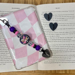 May include: A phone charm featuring pink, purple, black, and clear beads. The charm includes a circular charm with the text "Spook Babe" and heart-shaped charms.