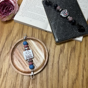 May include: A beaded keychain with a white rectangular charm that reads "Let's turn the page" with a floral design. The keychain has blue, pink, and patterned beads, and silver-colored clasps. The keychain is on a wooden plate.