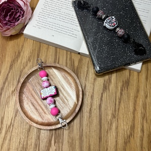 May include: A beaded keychain in pink and white, with a lobster clasp. It has a charm that says "My Book Mood Is Not Mentally". The keychain is on a wooden tray, beside a phone case and a book.