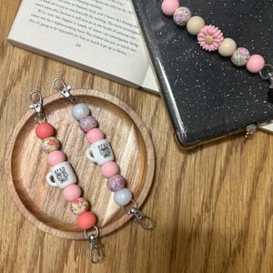 May include: Two beaded lanyards with lobster claw clasps. One lanyard features pink, floral, and white beads, with a small mug that says "READ BOOKS." The other lanyard has similar beads and a mug that says "READ BOOKS."