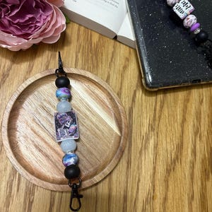 May include: A black phone charm with a white and purple beaded chain. The charm features a tarot card with a skeleton holding a book. The card reads "The Death".