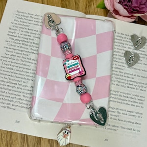 Op de afbeelding: Een doorzichtige telefoonhoes met een roze en wit geblokt patroon, versierd met een kralenbedel. De bedel heeft roze kralen, bloemaccenten en een roze rechthoek met de tekst "This Kindle Contains Explicit Material."