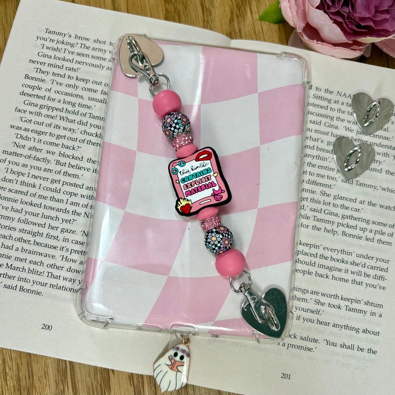 Op de afbeelding: Een doorzichtige telefoonhoes met een roze en wit geblokt patroon, versierd met een kralenbedel. De bedel heeft roze kralen, bloemaccenten en een roze rechthoek met de tekst "This Kindle Contains Explicit Material."