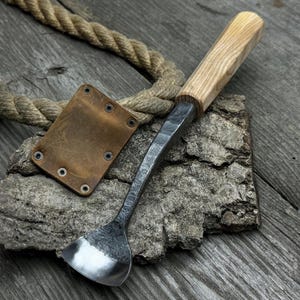 May include: A hand-forged spoon with a wooden handle, resting on a piece of bark. The spoon has a rounded bowl and a slightly curved handle. The handle is made of light-colored wood with a natural grain pattern. The spoon is a rustic and functional piece of handcrafted kitchenware.
