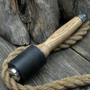 Puede incluir: Un mazo de madera con una cabeza de goma negra y un mango de madera natural. El mazo está sobre una cuerda, que está sobre una superficie de madera.