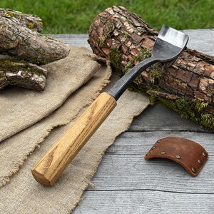 May include: A hand-forged metal drawknife featuring a polished, curved blade and a long, straight wooden handle with a visible grain. A separate, small, curved brown leather guard with metal rivets is positioned nearby on the surface.