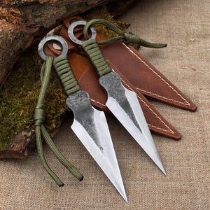 May include: Two forged steel throwing knives with sharp, triangular blades. The handles are wrapped in green cord, and each knife has a ring at the end. They rest on a brown leather sheath, with a natural wood and moss background.