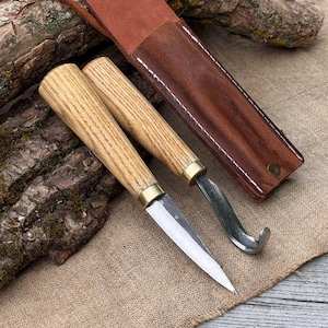 Puede incluir: Dos cuchillos para tallar madera con mangos de madera marrón claro y detalles de latón, junto con una funda de cuero marrón. Un cuchillo tiene una hoja recta, el otro una hoja curva. Herramientas para tallar madera.