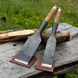 Peut inclure: Deux outils à main dotés de larges lames plates en métal gris foncé et de manches en bois brun clair. Chaque outil présente une virole en métal argenté près du manche et repose sur un petit étui carré en cuir brun protégeant le tranchant.