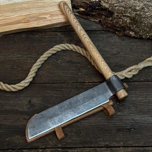 May include: A large, hand-forged cleaver with a wooden handle and a leather sheath. The blade is made of steel and has a rustic, worn appearance. The handle is light-colored wood. A rope is coiled around the cleaver.