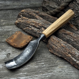 May include: A curved wood carving tool with a wooden handle and a metal blade. The tool is resting on a piece of wood with a leather patch nearby.