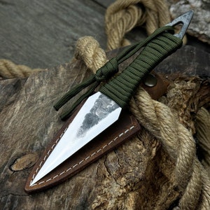Könnte beinhalten: Ein kleines, silbernes Messer mit einer grünen Paracord-Umwicklung und einer braunen Lederscheide. Das Messer liegt auf einem Stück Holz mit einem umwickelten Seil.