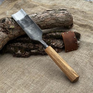 May include: A metal gouge with a curved, dark steel blade and a straight, light-colored wooden handle. Beside the tool lies a small, rectangular brown leather sheath secured with five metal rivets, designed to protect the sharp edge.