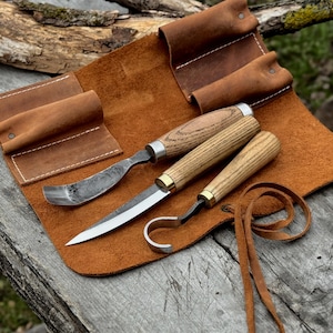 May include: A set of three wood carving tools with light-colored wooden handles and metal blades, including a curved gouge, a straight detail knife, and a hook knife, resting on a brown leather tool roll with stitched pockets and leather ties.