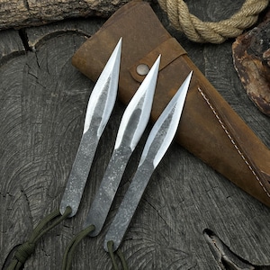 May include: Three silver throwing knives with brown leather sheaths. The knives are arranged in a fan shape on a dark wooden surface.