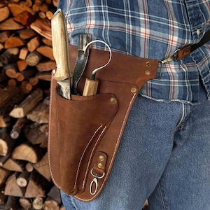 May include: A brown leather knife sheath with three knives and a hook. The sheath is worn on the waist and has white stitching and metal rivets. The background is a stack of firewood.