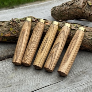 May include: Five wooden handles with brass collars are displayed on a weathered wooden surface. The handles have a natural wood grain pattern and a cylindrical shape. A piece of wood rests in the background.