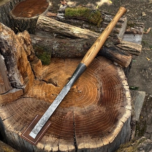 Peut inclure: Un ciseau à bois à long manche avec une lame en acier gris foncé texturé et un manche en bois brun clair présentant un motif de grain prononcé. L'extrémité tranchante du ciseau est protégée par un petit étui en cuir brun rectangulaire.