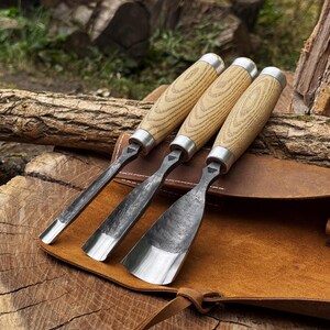 May include: Three wood carving chisels with light-coloured wooden handles and silver metal ferrules. The blades are made of dark, forged steel with varying widths, including a narrow gouge, a medium gouge, and a wide, flat-edged chisel blade.