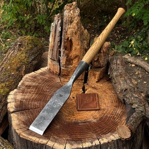 May include: A hand-forged steel chisel with a long, light-brown wooden handle featuring a visible grain. The metal blade has a dark, textured finish and a small stamped logo. A small, square brown leather patch with a logo is placed nearby.
