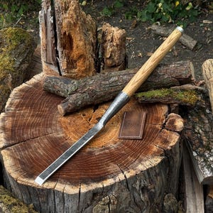 May include: A long, narrow metal chisel with a straight, flat cutting edge and a cylindrical wooden handle featuring a visible grain pattern. Beside the chisel lies a small, rectangular brown leather sheath embossed with a geometric logo.