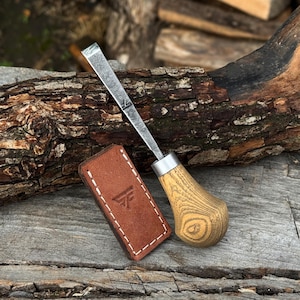 Peut inclure: Un ciseau à bois en métal avec un manche en bois brun clair et une virole en métal argenté. La lame du ciseau présente un petit logo gravé. À côté de l'outil se trouve un étui rectangulaire en cuir brun avec des coutures blanches.