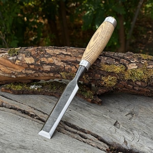 Peut inclure: Un ciseau à bois doté d'une lame en acier plate et rectangulaire avec un petit logo estampillé, ainsi qu'un manche en bois clair texturé muni d'une virole et d'un embout en métal argenté, conçu pour le travail du bois et la sculpture.