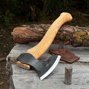 Può includere: Un'accetta con manico in legno chiaro curvato e testa in metallo forgiato scuro con un bordo affilato e lucido. Un piccolo fodero in pelle marrone con un bottone a pressione in metallo è posizionato accanto alla testa dell'ascia su legno.