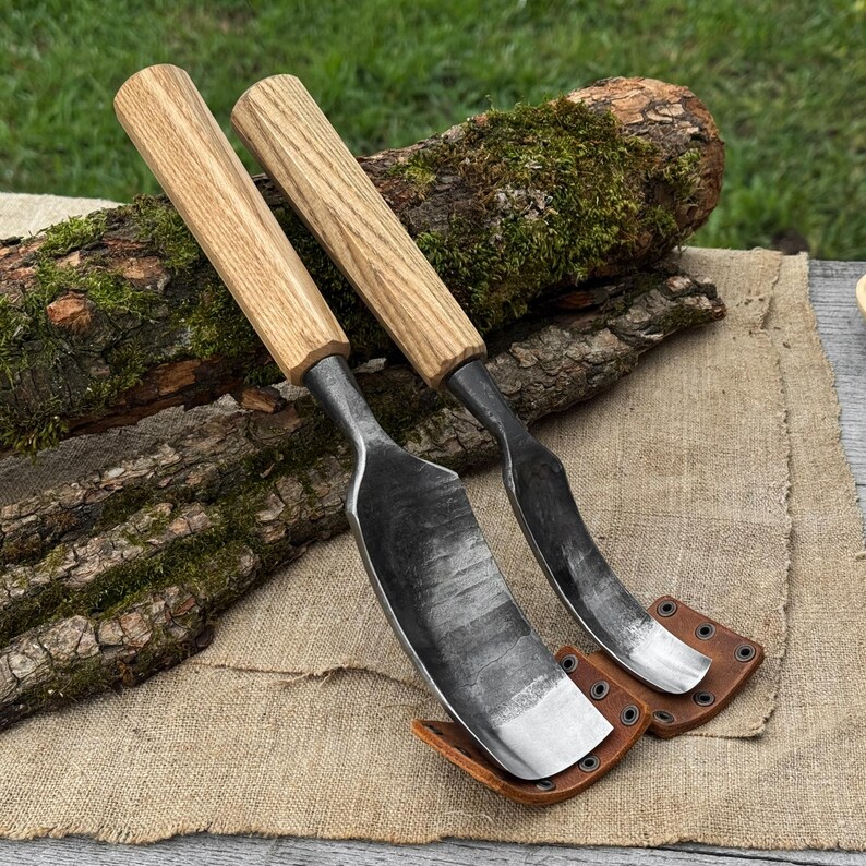 May include: Two hand-forged draw knives with light wood handles and dark metal blades. The blades are curved and have a sharp edge. Each knife rests in a brown leather sheath with rivets. The knives are displayed on a burlap surface.