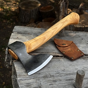 May include: A hand axe with a curved light brown wooden handle and a dark forged metal head featuring a sharp, polished edge. A brown leather sheath with a snap button closure and a stamped logo is positioned next to the axe head.