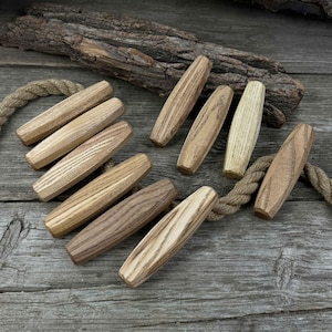 May include: A collection of nine elongated, spindle-shaped wooden cabinet pulls with visible natural grain patterns. Each handle features a smooth, tapered design with a light to medium brown finish, arranged in a cluster on a textured surface.