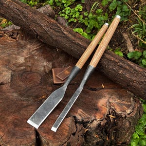 Peut inclure: Deux ciseaux à bois dotés de longs manches en bois clair et de lames en métal forgé sombre. Les lames présentent une finition martelée texturée et un tranchant biseauté. Un petit étui en cuir marron de forme carrée est placé près des manches.