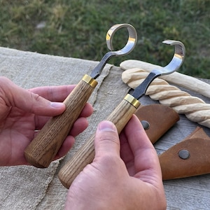 Könnte beinhalten: Zwei Holzwerkzeuge mit gebogenen Stahlklingen und Holzgriffen. Die Griffe sind hellbraun mit einem messingfarbenen Band am Klingensockel. Ein Werkzeug hat eine ringförmige Klinge, das andere eine hakenförmige Klinge.