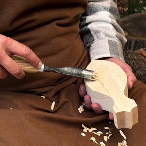 May include: A metal wood carving gouge with a light-colored wooden handle is being used to carve a piece of light-colored, unfinished wood. The wood is shaped with a pointed end and a wider, rounded body, showing visible wood shavings.