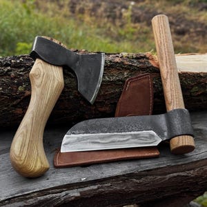 May include: Two axes and a cleaver with wooden handles and dark metal blades rest on a wooden surface. The cleaver has a leather sheath. The axes have a classic design, while the cleaver has a wide, curved blade. The tools are likely for woodworking or outdoor use.