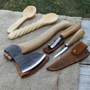 Puede incluir: Una colección de herramientas de carpintería hechas a mano, que incluye dos cucharas de madera, un hacha con funda de cuero y dos cuchillos con fundas de cuero. Las herramientas están dispuestas sobre una tela de tono neutro.