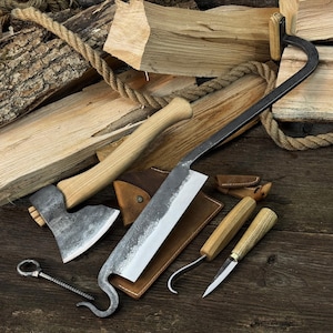 Puede incluir: Una colección de herramientas rústicas forjadas a mano, que incluyen un hacha, un cuchillo grande y cuchillos más pequeños con mangos de madera. Las herramientas están dispuestas sobre una superficie de madera con leña y cuerda de fondo, mostrando una estética tradicional y utilitaria.