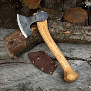 May include: A hand axe with a dark, forged steel head featuring a small triangular logo and a curved, light-colored wooden handle with prominent grain patterns. A brown leather sheath with brass rivets and a matching logo is positioned nearby.