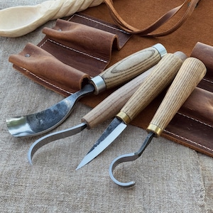 Puede incluir: Un juego de cuatro herramientas para tallar madera con mangos de madera y hojas de metal, exhibidas con un estuche de cuero. Las herramientas tienen diferentes formas de hoja, incluyendo una gubia curva, un cuchillo recto y un cuchillo de gancho.