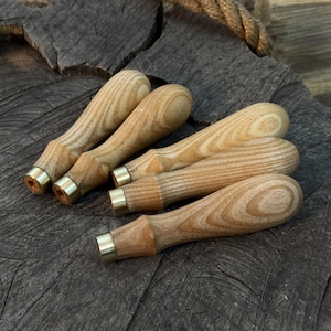 以下が含まれることがあります： 真鍮のフェルールが付いた、木製のノミの柄が5つ。柄は木目がはっきり見える明るい色の木材で作られています。真鍮のフェルールが工具に金属的なアクセントを加えています。ノミは風化した木製の表面に配置されています。