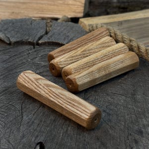 Puede incluir: Cinco espigas de madera con un acabado marrón claro y veta de madera visible están dispuestas sobre una superficie oscura y texturizada. Las espigas son cilíndricas con un extremo hexagonal y un pequeño agujero. Los tonos de madera natural crean una estética rústica.