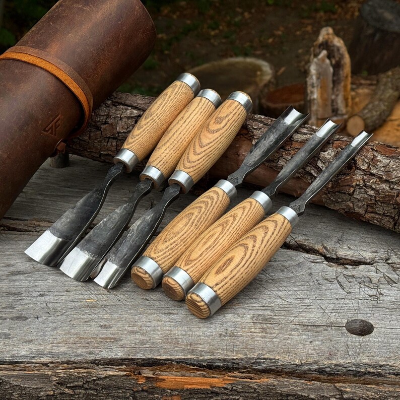 May include: A set of six wood carving gouges with light brown wooden handles and silver metal ferrules. The tools feature curved, dark grey steel blades of varying widths. A brown leather tool roll is positioned to the left of the gouges.