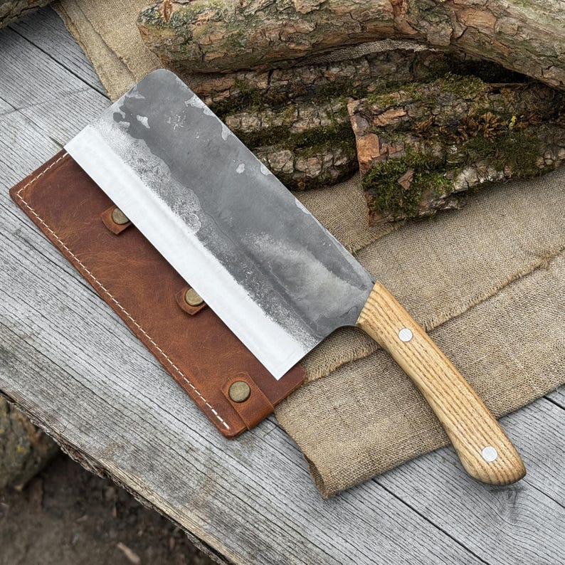 Puede incluir: Una gran cuchilla r&uacute;stica con un mango de madera clara y una hoja ancha y gris. La cuchilla descansa sobre una superficie de madera junto a una funda de cuero marr&oacute;n. La hoja mide unos 30 cm de largo.