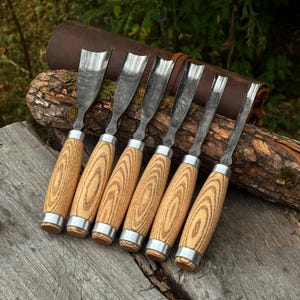 May include: A set of six wood carving gouges with curved metal blades and light brown wooden handles featuring a prominent grain pattern. Each tool has a silver metal ferrule connecting the blade to the handle, arranged in a row.