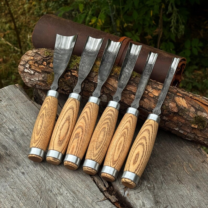 May include: A set of six wood carving gouges with curved metal blades and light brown wooden handles featuring a prominent grain pattern. Each tool has a silver metal ferrule connecting the blade to the handle, arranged in a row.