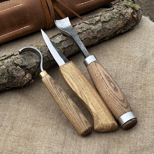 May include: A set of three wood carving tools with wooden handles and metal blades. The tools include a hook knife, a straight knife, and a curved chisel. The handles are light brown, and the blades are silver. A brown leather case is visible in the background.