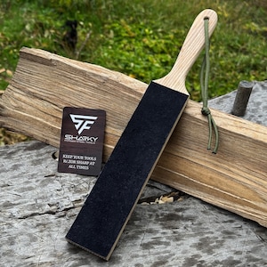 May include: A wooden razor strop with a black leather surface and a light-colored wooden handle. The strop is displayed with a business card that reads "SHARKY FORGED STEEL TOOLS" and "KEEP YOUR TOOLS RAZOR SHARP AT ALL TIMES."