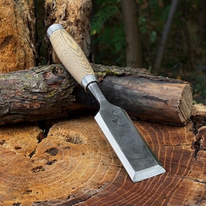 May include: A woodworking chisel featuring a wide, flat, dark steel blade with a small stamped logo and a light-colored, textured wooden handle with a metal ferrule. The tool is positioned diagonally across a rough, circular cut log section.