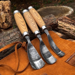 May include: Three wood carving tools with light-colored wooden handles and silver metal ferrules. Each tool features a curved, dark, forged steel blade with a textured finish and a small stamped logo near the handle connection point.
