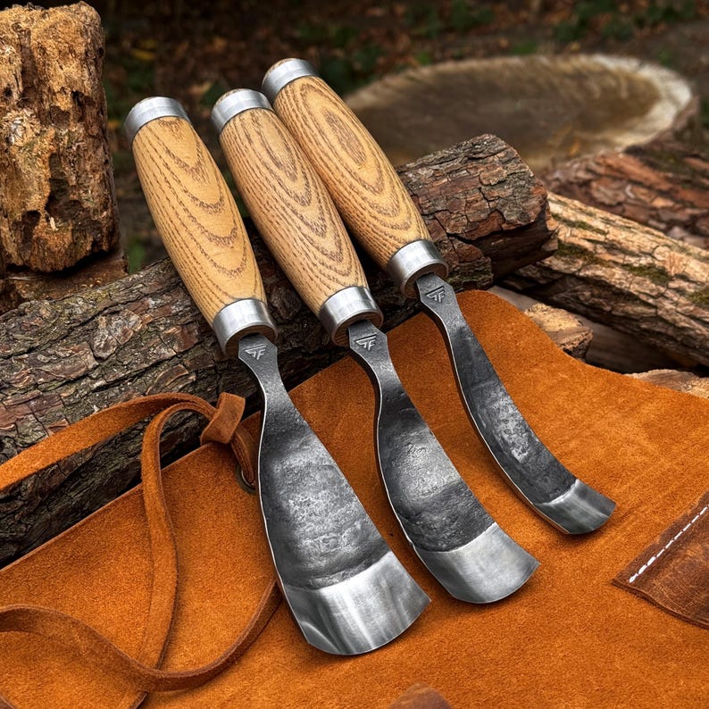 Puede incluir: Tres herramientas para tallar madera con mangos de madera clara y virolas de metal plateado. Cada herramienta presenta una hoja curva de acero forjado oscuro con acabado texturizado y un peque&ntilde;o logotipo estampado cerca del mango.